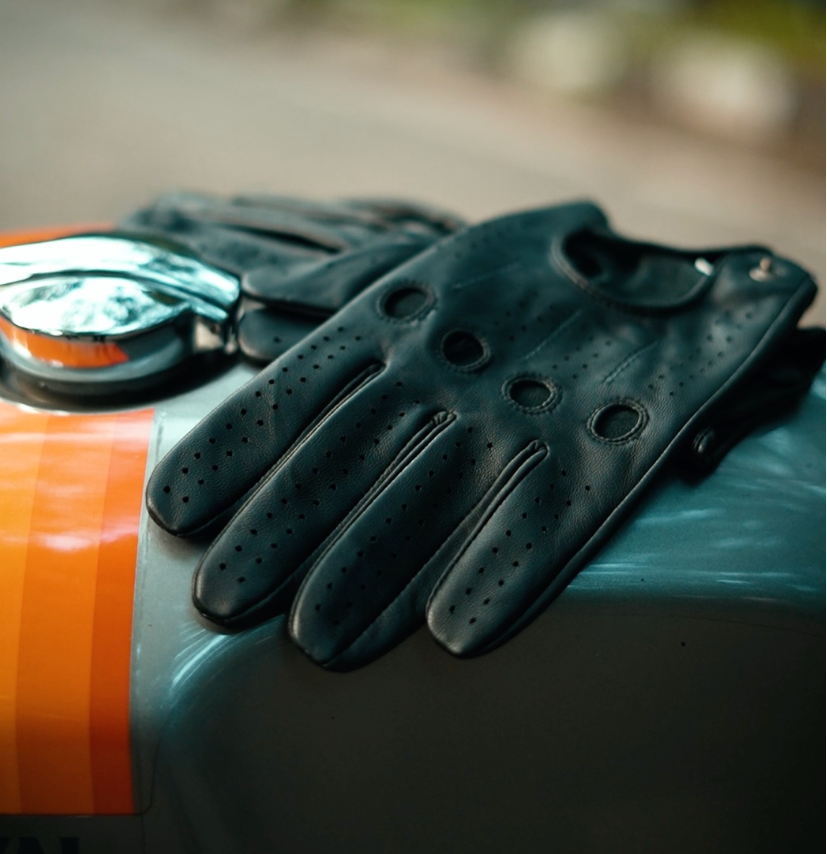 Black leather motorcycle gloves with perforated detailing, resting on the tank of a motorcycle with an orange stripe.