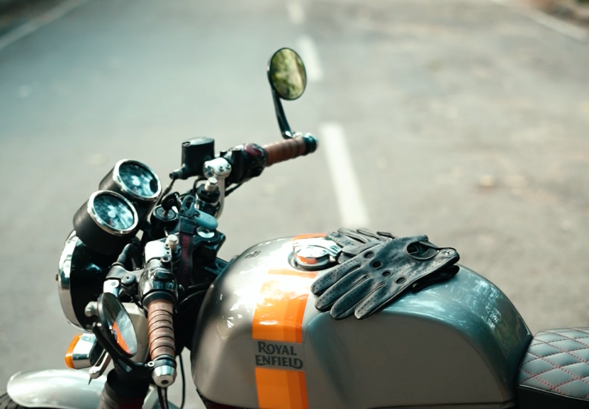 A close-up shot of a motorcycle tank and handlebar area on an asphalt road. The tank is dark gray with a bold orange stripe and the words 'ROYAL ENFIELD' printed on it.